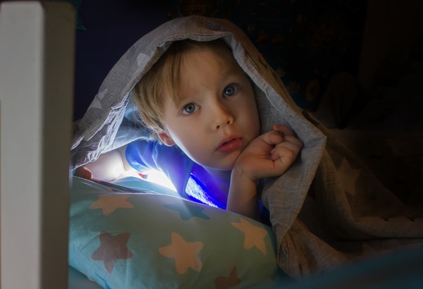 Child lying in bed with covers over their head looking at a phone.